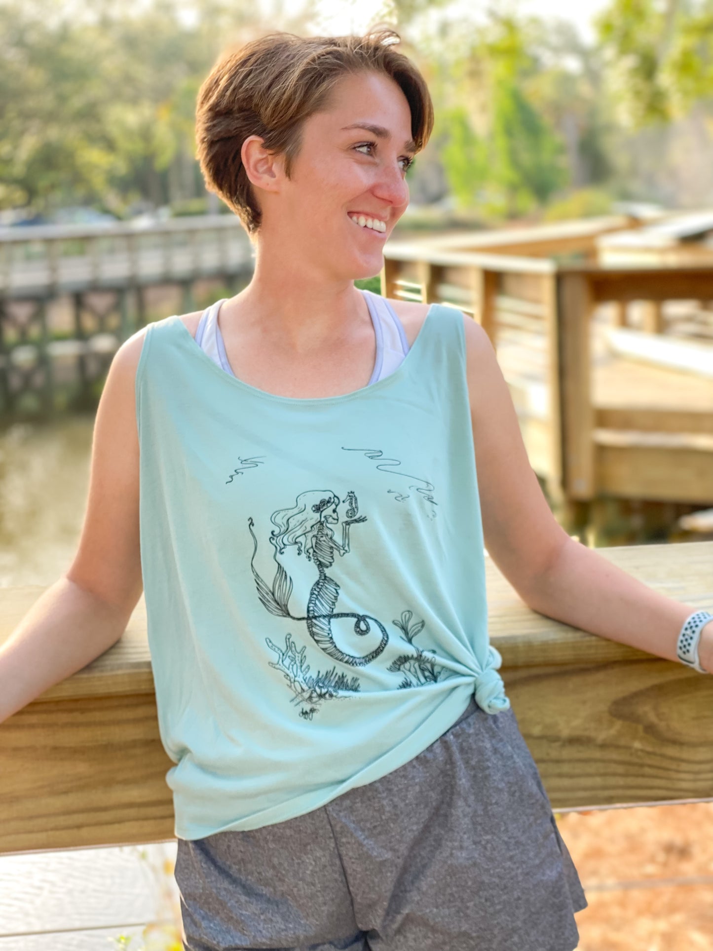 Smiling girl wearing a dusty blue tank top featuring a black skeleton mermaid design